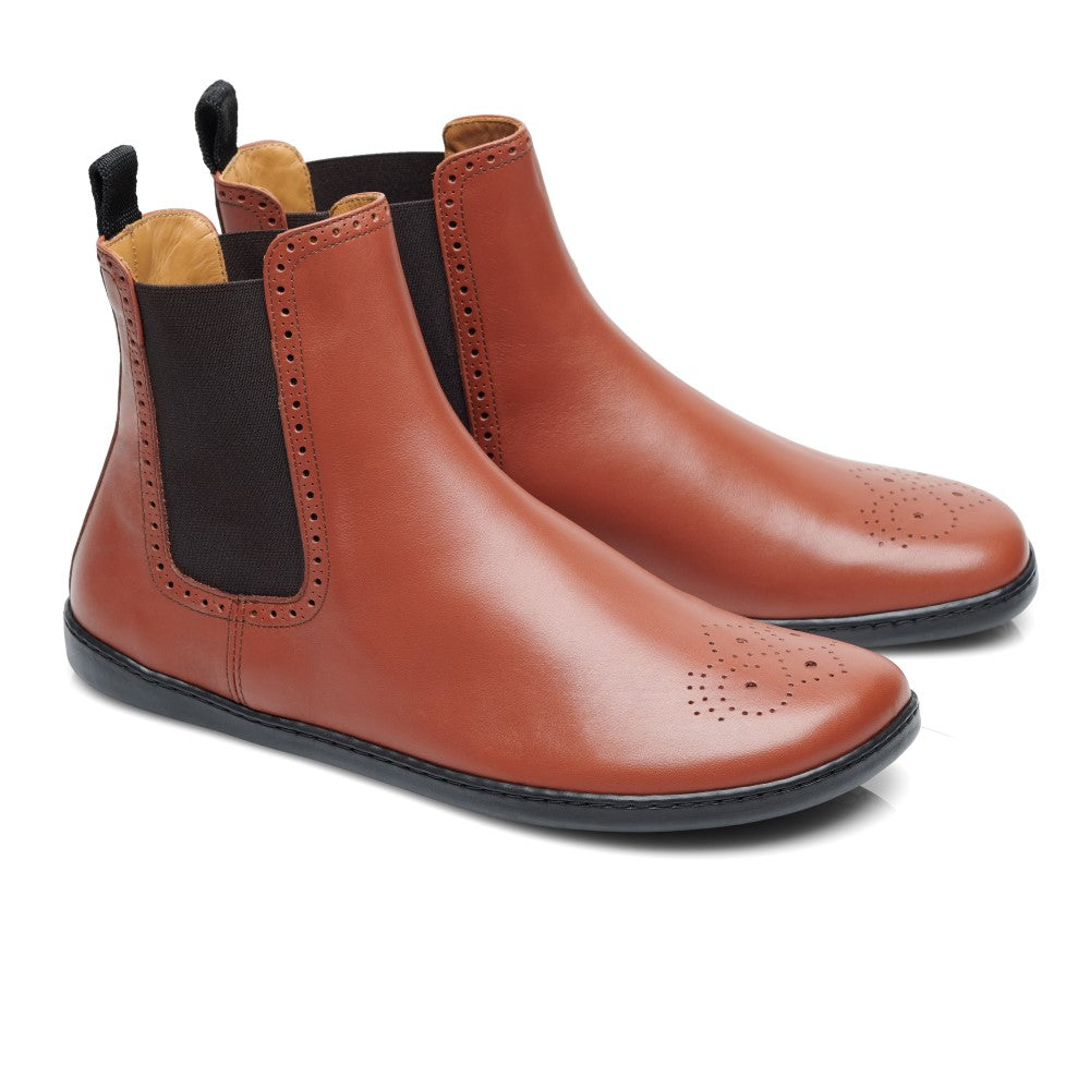 Die EQUITY Brogue Cognac-Stiefel von ZAQQ verfügen über schwarze elastische Seitenteile und aufwendige Brogue-Perforationen an der Spitze. Ihre schlichte Ästhetik wird durch ein hellbraunes Innenfutter unterstrichen, während sich das Mid-Cut-Design elegant von einem weißen Hintergrund abhebt.