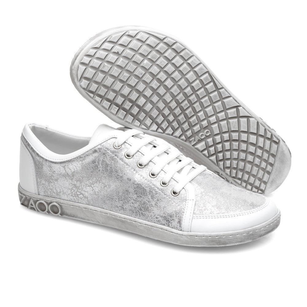 Die TIQQ Silver White Sneaker von ZAQQ zeichnen sich durch ein stilvolles Design mit einem strukturierten grauen Muster auf der weißen Oberfläche aus. Ein Sneaker steht aufrecht und zeigt die sichtbaren Schnürsenkel, während der andere die markante Sohle mit Waffelmuster zeigt. Sie sind aus hochwertigem Nappaleder gefertigt und zeigen stolz das ZAQQ-Logo an der Seite der Sohle.